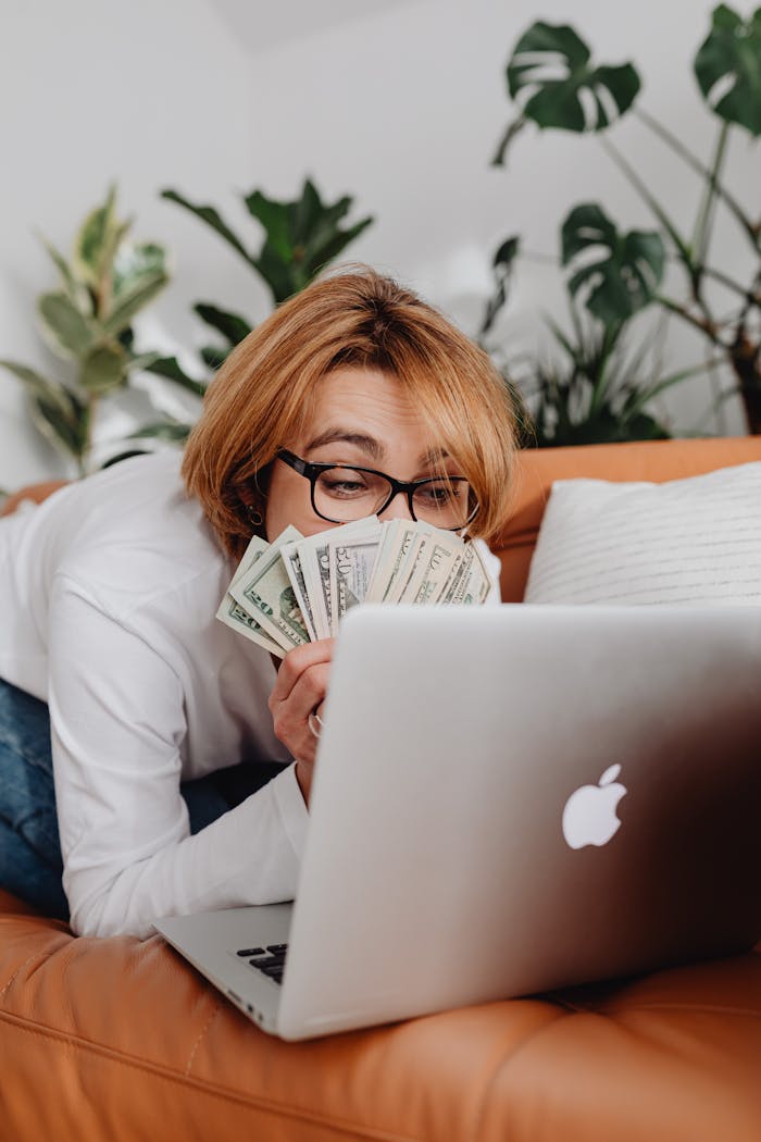 Woman lying on couch using laptop with cash in hand, surrounded by houseplants.